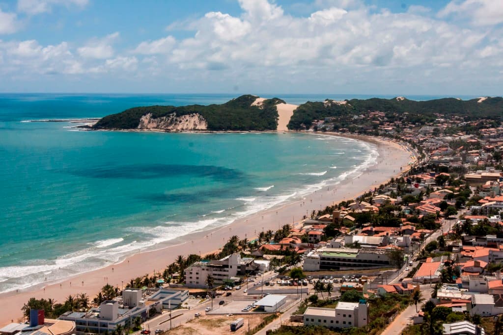 Praia de Ponta Negra em Natal com areia dourada, mar azul, coqueiros e o Morro do Careca ao fundo.